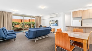A modern hotel room with a sofa, a dining table with chairs, a TV, and a kitchen area with cabinets. - APX Parramatta (Rosehill)