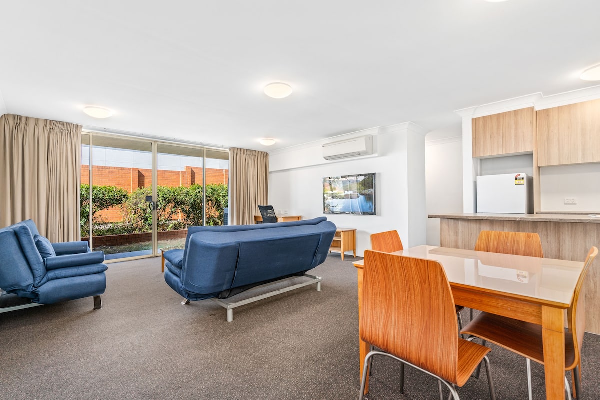 A modern hotel room with a sofa, a dining table with chairs, a TV, and a kitchen area with cabinets.