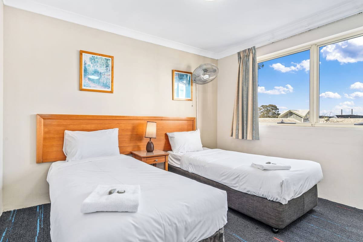 A hotel room with two beds, a wooden headboard, a bedside table with a lamp, a window with curtains, and two framed pictures on the wall.