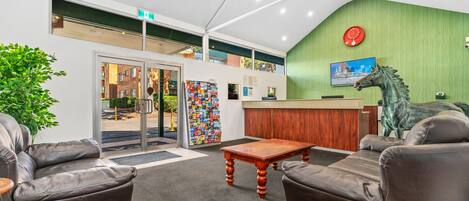 A lobby with leather sofas, a wooden coffee table, a reception desk, a green wall with a clock, and a cityscape painting.