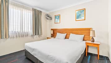A hotel room with a large bed, a wooden headboard, a bedside table with a lamp, a wall-mounted fan, and two framed pictures on the wall.