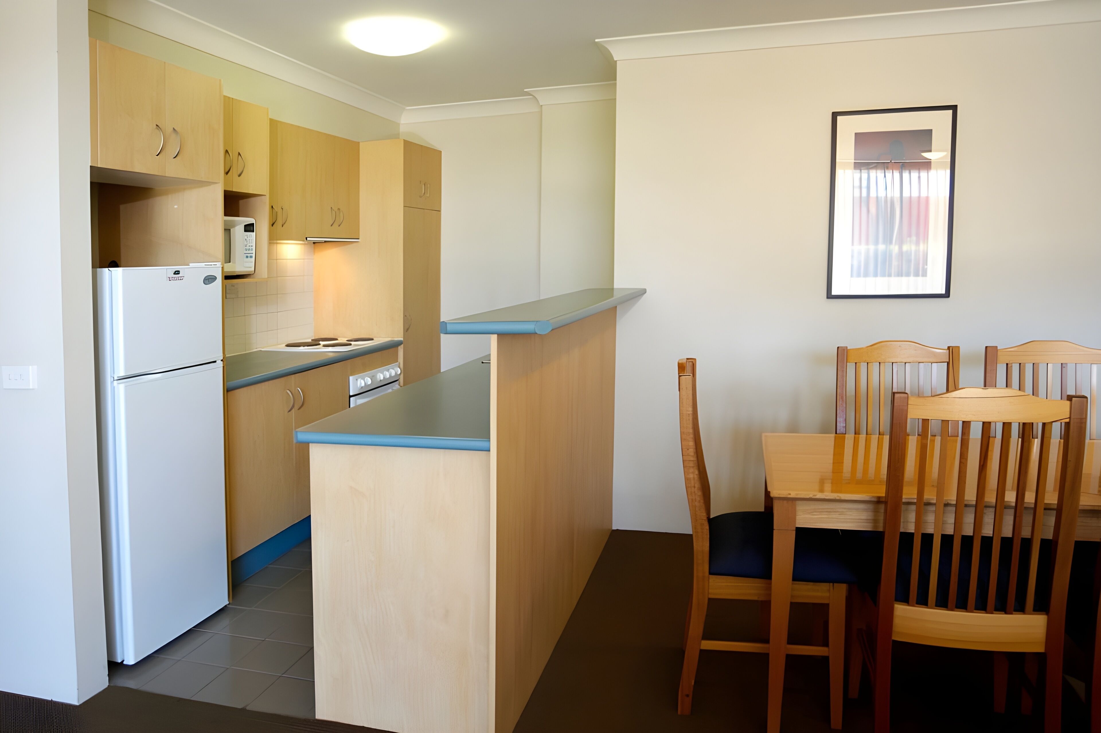 A compact kitchen with wooden cabinets, a white refrigerator, a microwave, and a small dining area with a table and chairs.