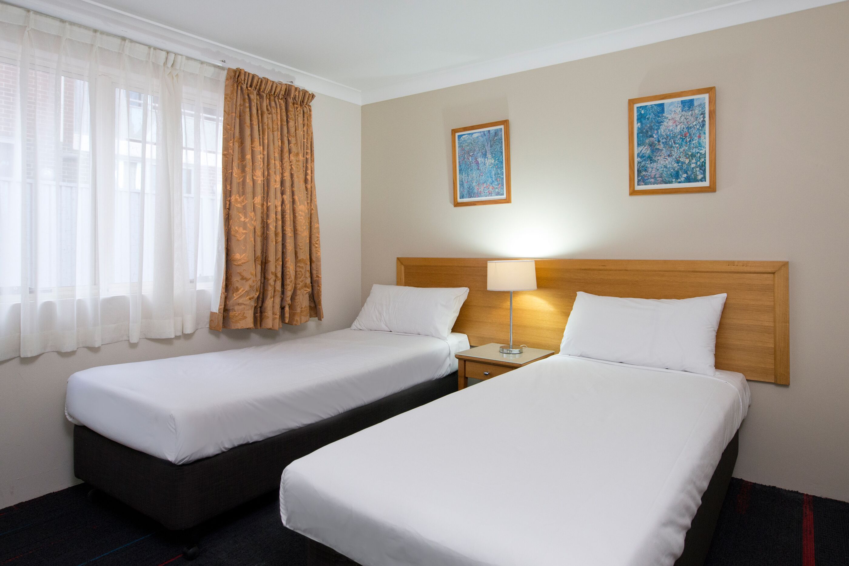 Habitación de hotel con dos camas individuales, una mesita de noche con lámpara y dos cuadros enmarcados en la pared.