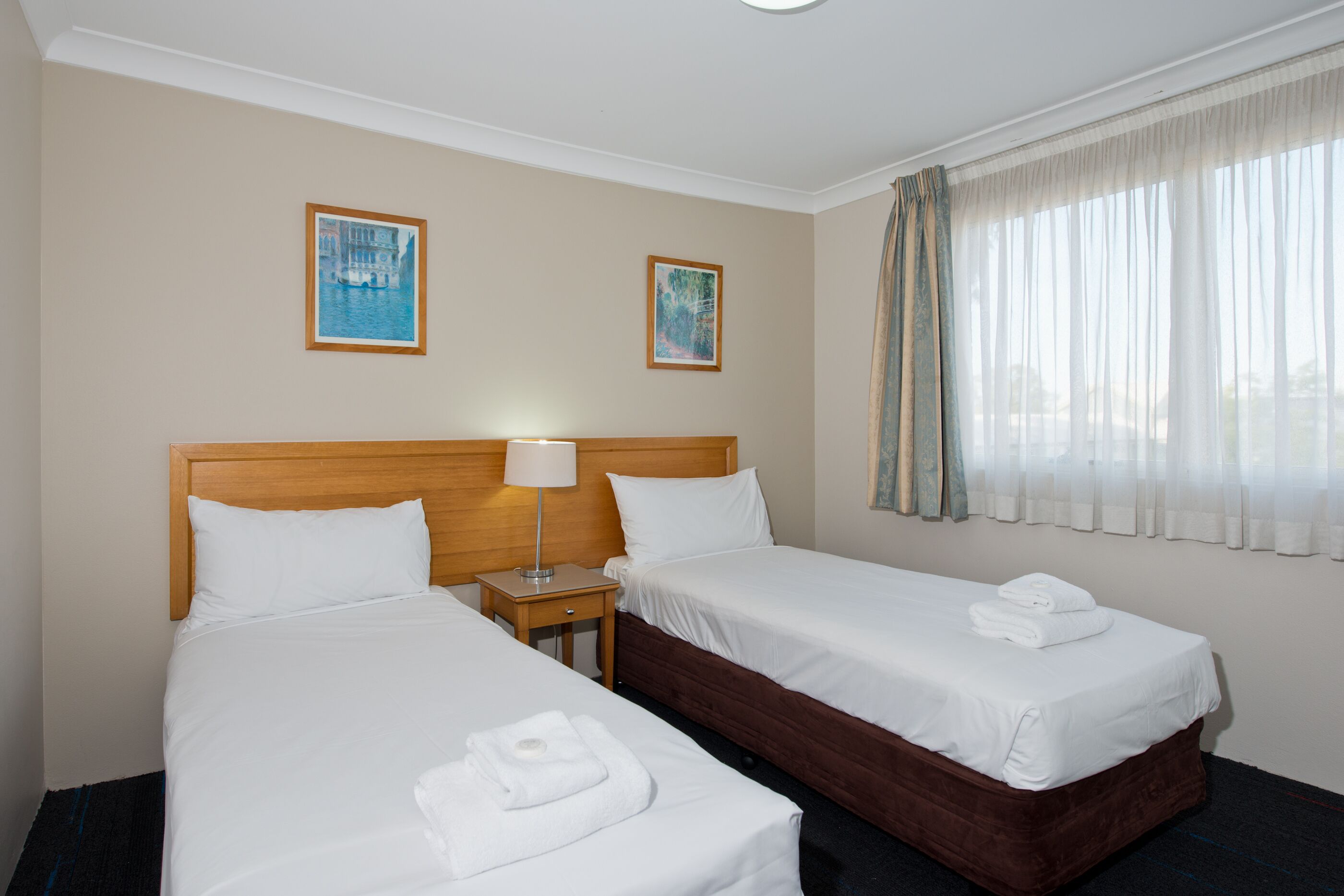 Habitación de hotel con dos camas individuales, cabecera de madera, mesita de noche con lámpara y dos cuadros en la pared.