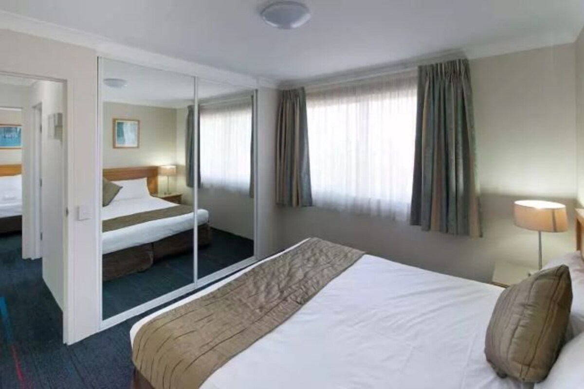 A hotel room with a large bed, a mirror, a nightstand, and a window with curtains.