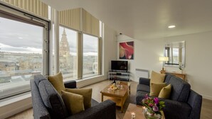 A modern living room with two sofas, a coffee table, and a TV. Large windows offer a city view. - PREMIER SUITES Liverpool (Liverpool)