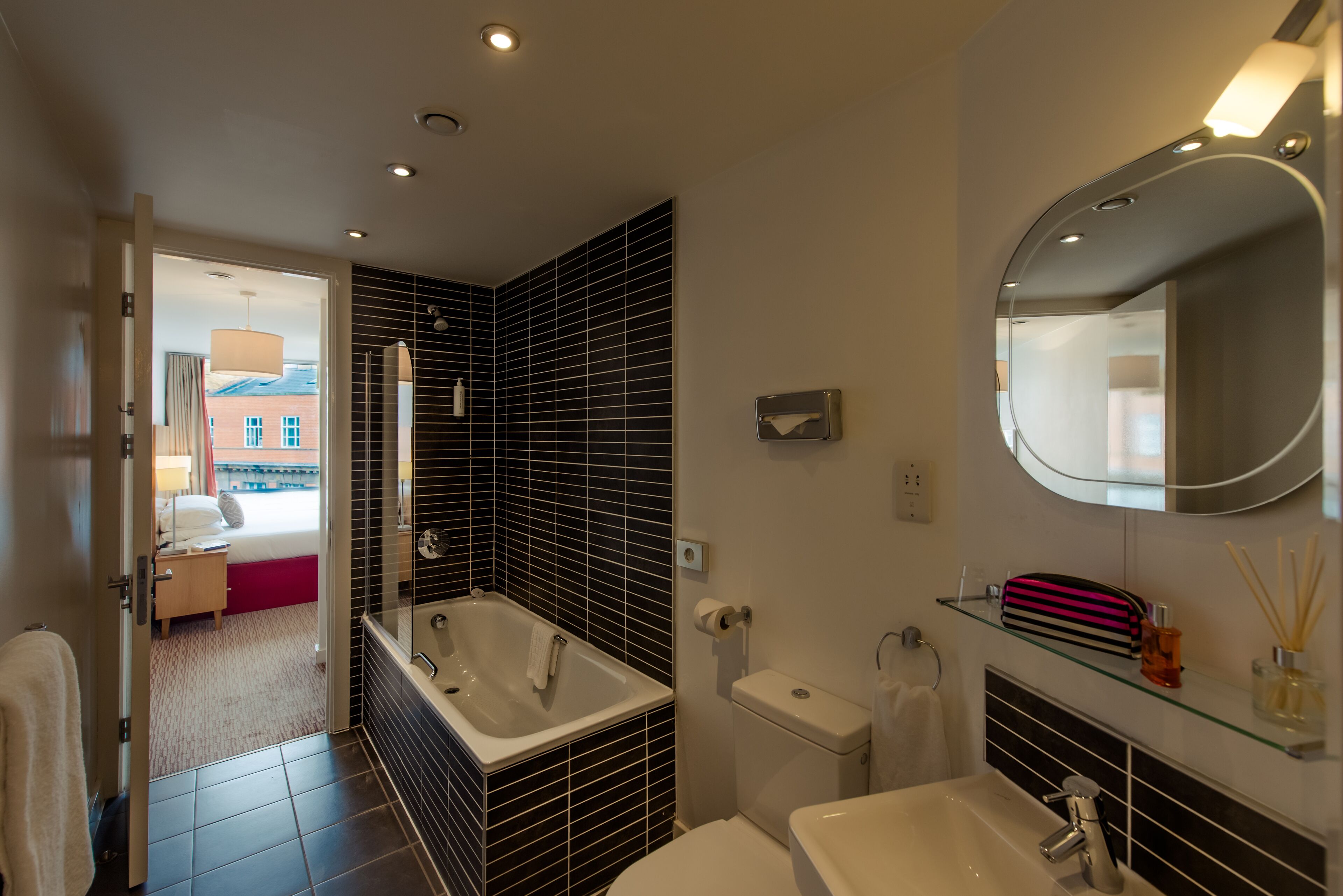 Ein modernes Badezimmer mit Badewanne, Toilette und Waschbecken. Über dem Waschbecken befindet sich ein Spiegel und durch eine geöffnete Tür ist eine Duschkabine zu sehen.