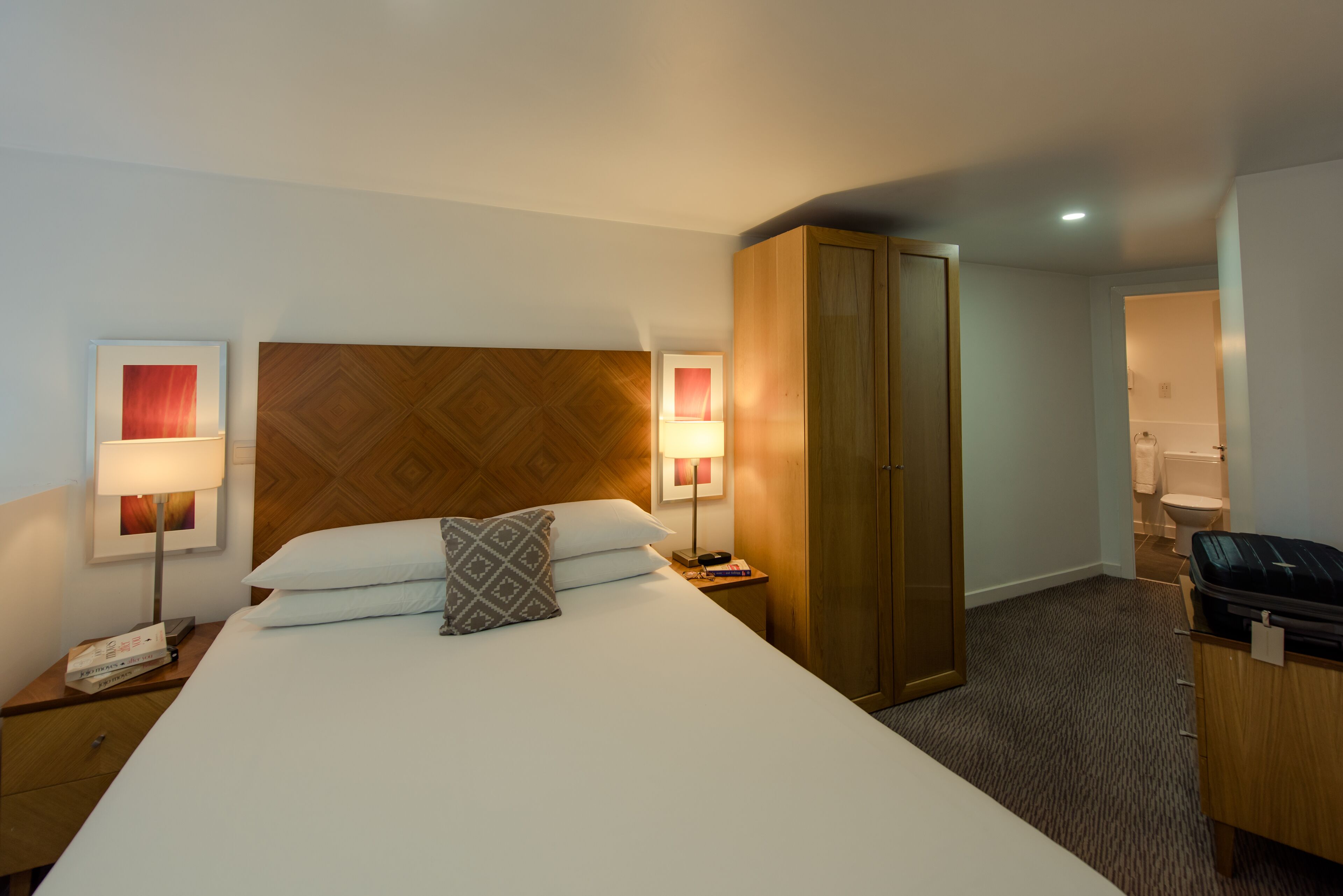 A hotel room with a bed, two bedside lamps, a wooden headboard, a wardrobe, a desk with a telephone, and a trash bin.