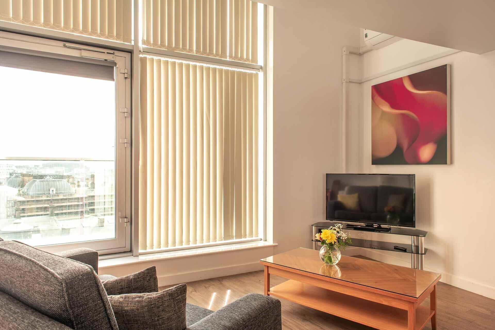 A modern living room with a sofa, a coffee table with a vase of flowers, a flat-screen TV, and a large window with blinds.