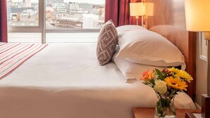 A hotel room with a large bed, a nightstand with a lamp, a vase with flowers, and a view of a cityscape with a tower. - PREMIER SUITES Liverpool (Liverpool)