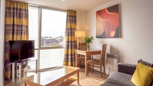 A modern living room with a flat-screen TV, a glass-top coffee table, a sofa, and a dining table with chairs. There is a painting on the wall and a window with curtains. - PREMIER SUITES Liverpool (Liverpool)