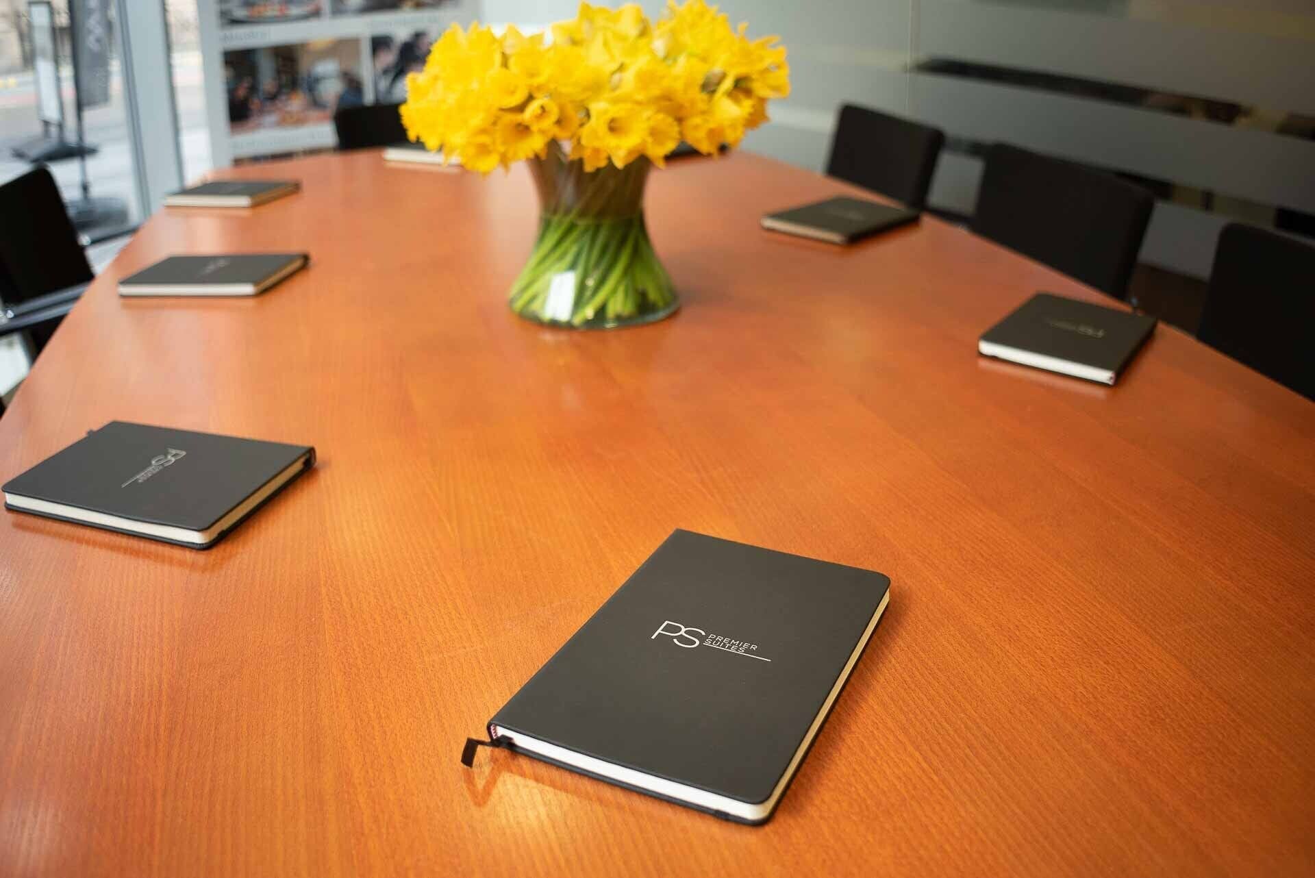 Ein Konferenztisch mit einem Blumenstrauß aus gelben Blüten, umgeben von vier schwarzen Notizbüchern.