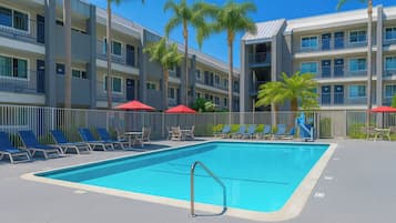 Une piscine entourée d’une clôture et de palmiers, avec des chaises longues et des parasols, et un immeuble à plusieurs étages en arrière-plan.