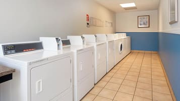 A laundry room with multiple white washing machines and dryers, a control panel, and a tiled floor.