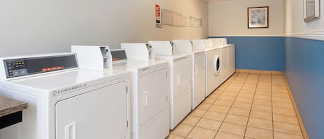 A laundry room with multiple white washing machines and dryers, a control panel, and a tiled floor.