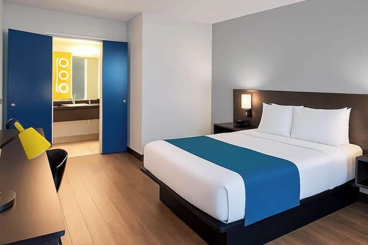 A hotel room with a bed, bedside tables, a desk with a chair, and a bathroom visible through an open door.