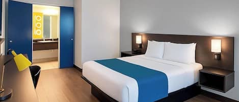 A hotel room with a bed, bedside tables, a desk with a chair, and a bathroom visible through an open door.