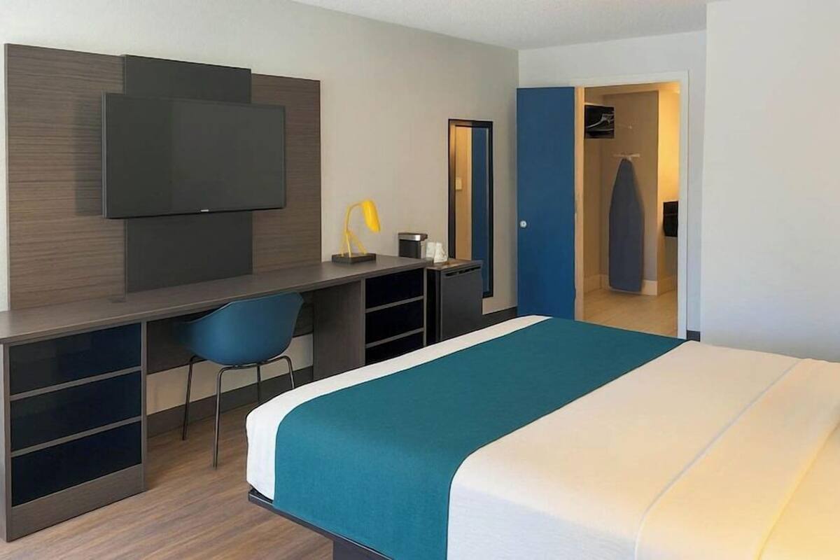 A modern hotel room with a bed, a desk, a chair, a television, and a wardrobe.