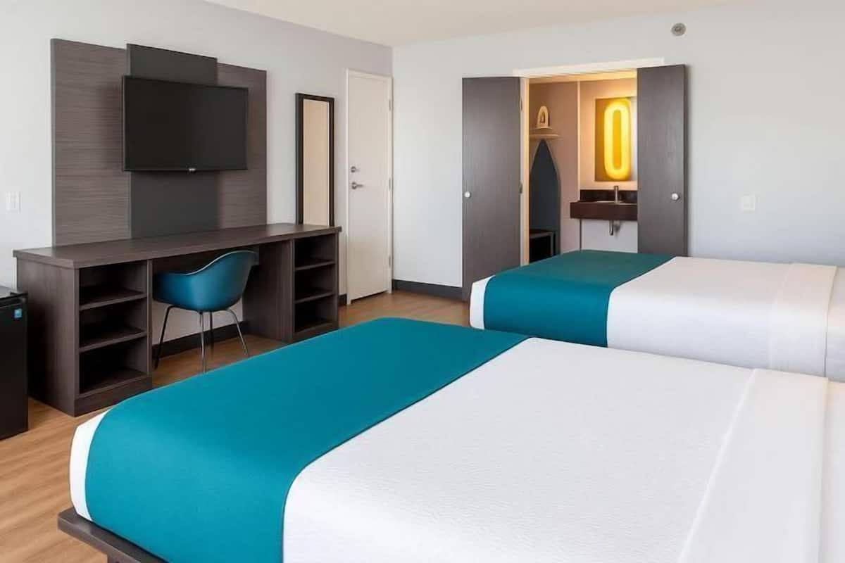 A hotel room with a desk, two beds, a TV, a chair, and a bathroom with a mirror and sink.