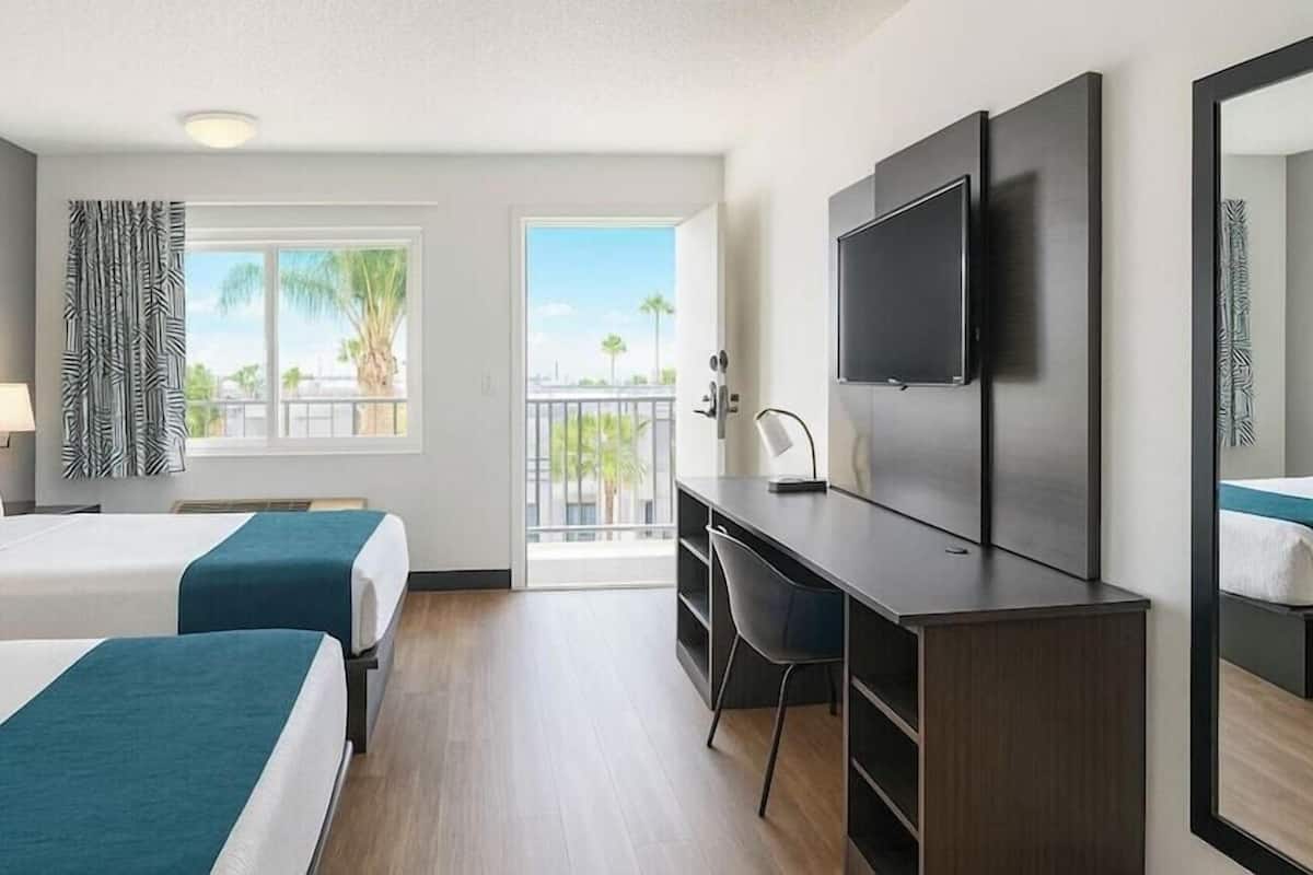A hotel room with two beds, a desk, a chair, a TV, and a balcony with a view of palm trees.
