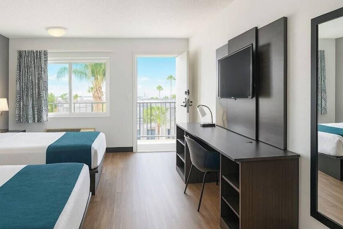 A hotel room with two beds, a desk, a chair, a TV, and a balcony with a view of palm trees.