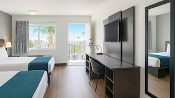 Habitación de hotel con dos camas, un escritorio, una silla, un televisor y un balcón con vista a palmeras.