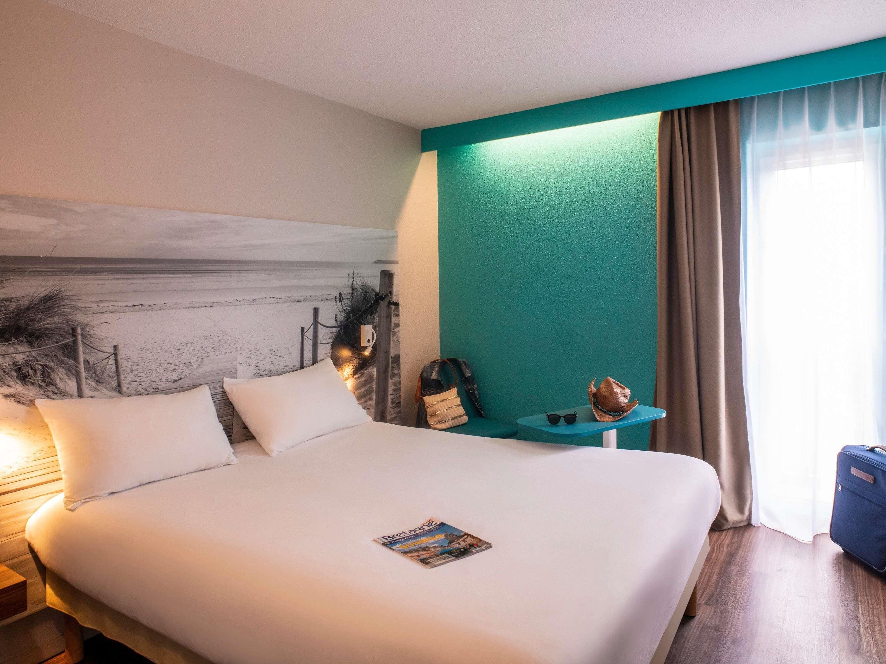 Habitación de hotel con una cama grande, una mesita con una revista, una maleta y vistas a la playa a través de la ventana.
