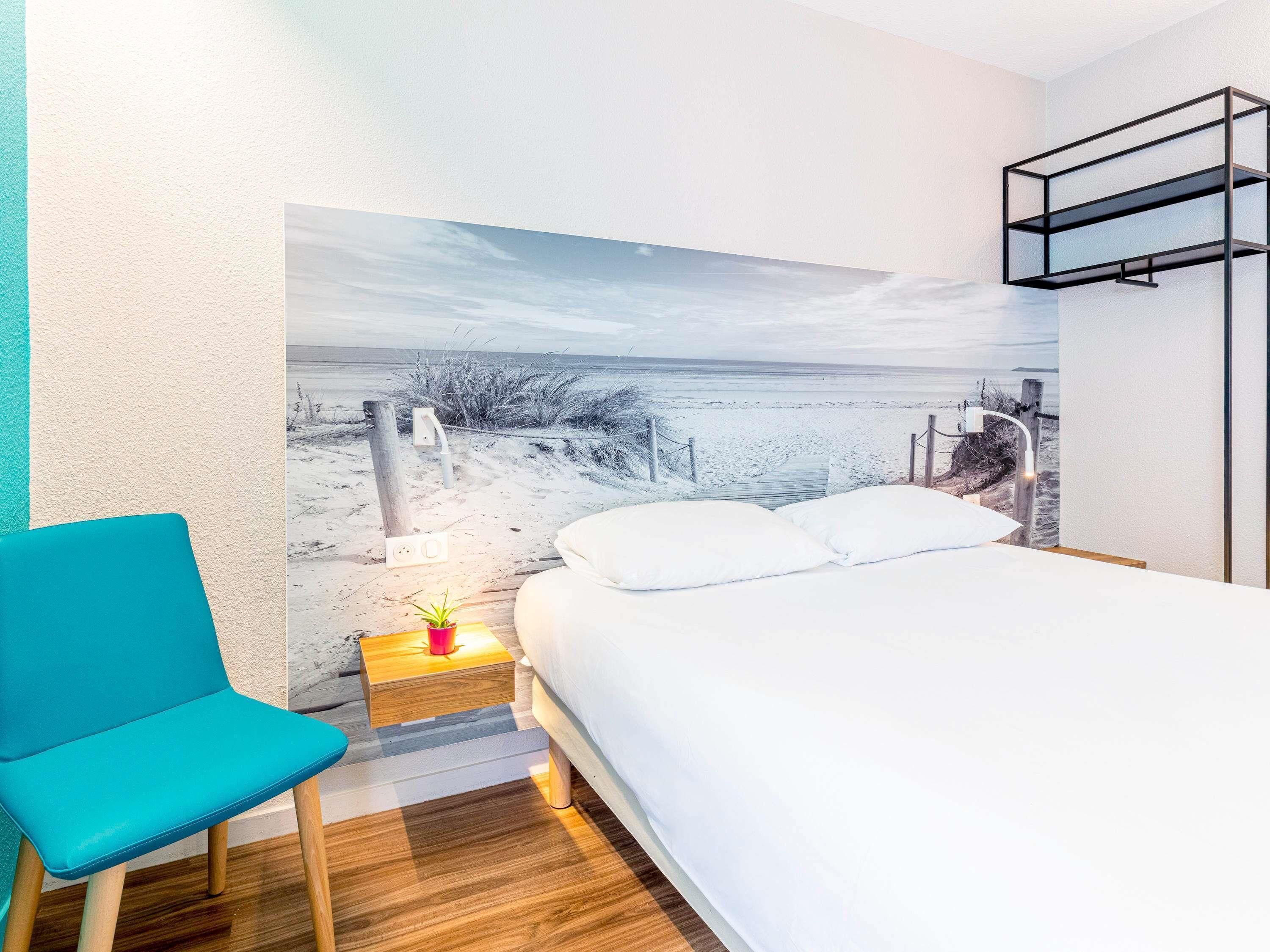 Un dormitorio con cama, una silla azul, una mesita de noche con lámpara y un mural en la pared que representa una playa.