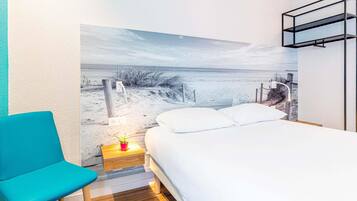 Un dormitorio con cama, una silla azul, una mesita de noche con lámpara y un mural en la pared que representa una playa.
