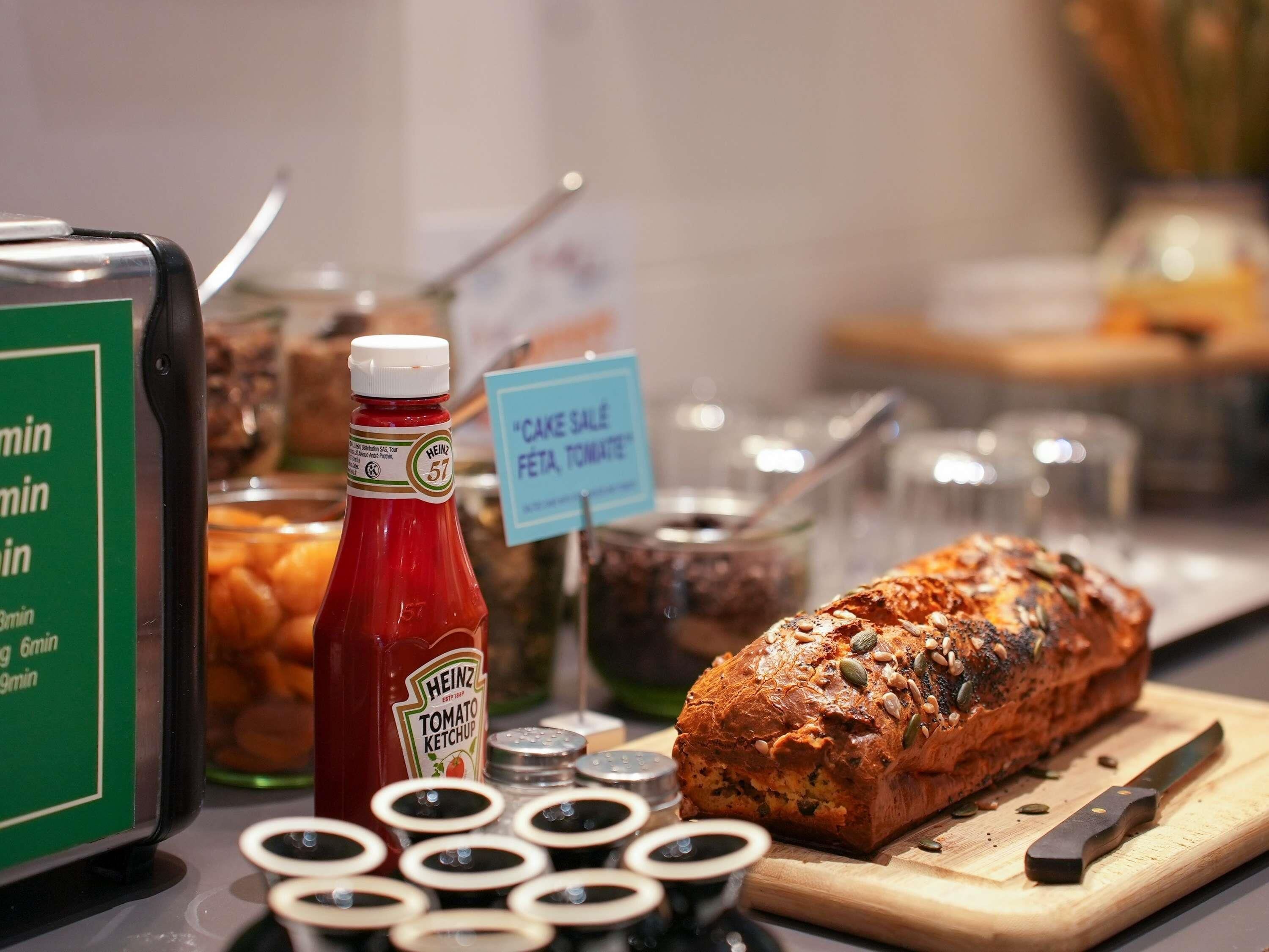 A table with a bottle of Heinz Tomato Ketchup, a loaf of seeded bread, condiments, and a sign indicating 'Cake Sale Peta, Tomate'.