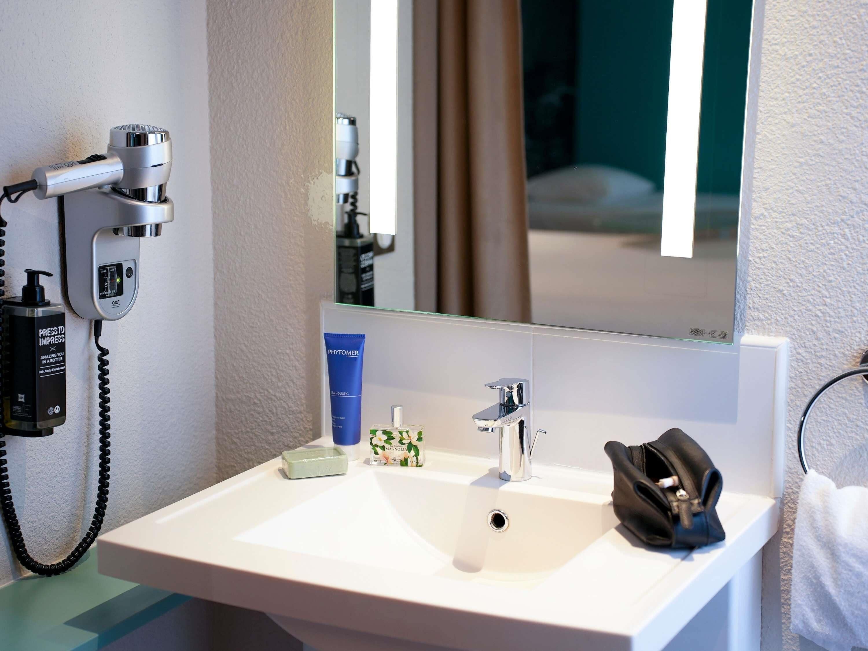 A hotel bathroom with a white sink, a mirror, a hairdryer, a bottle of hand soap, and a black bag on the counter.