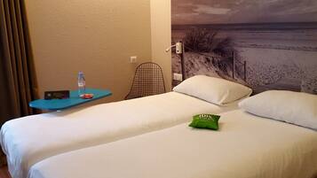 Quarto de hotel com uma cama, uma pequena mesa com uma garrafa de água, um travesseiro e uma etiqueta verde do hotel.