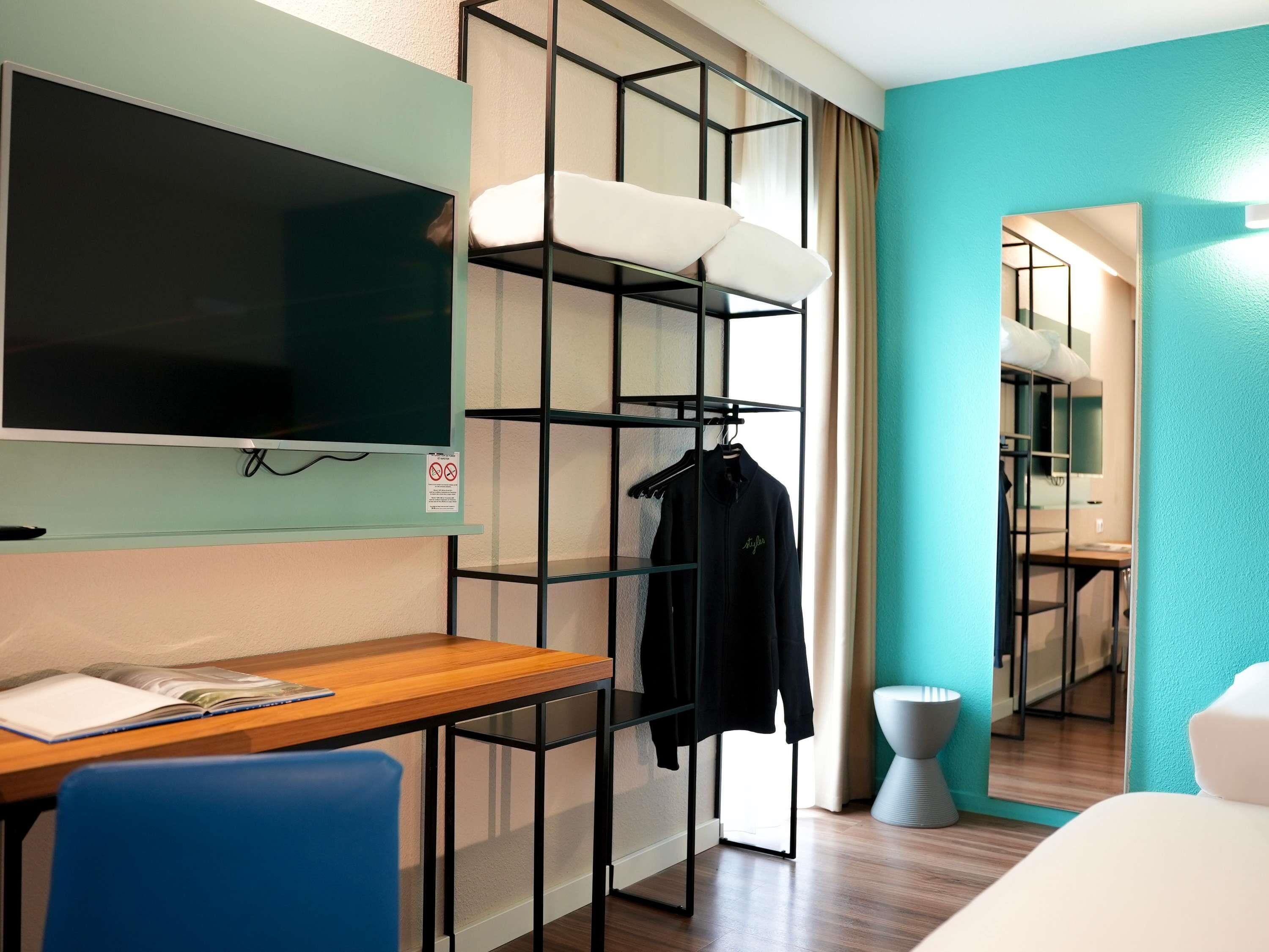 A hotel room with a wooden desk, a TV, a black metal shelf with clothes, a white chair, and a mirror.