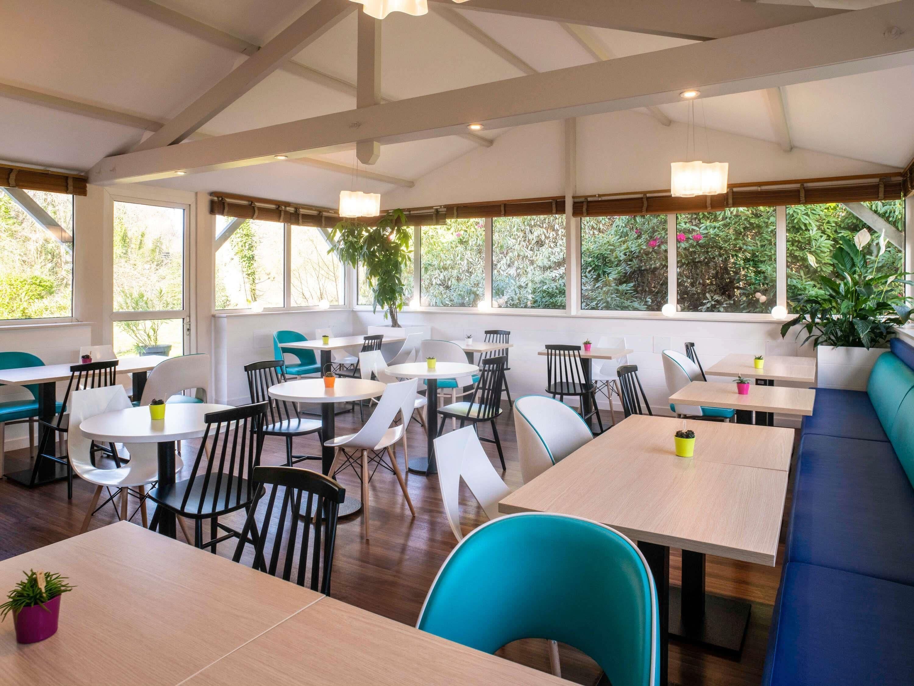 Un comedor moderno con varias mesas y sillas, ventanas amplias con luz natural y plantas de interior.
