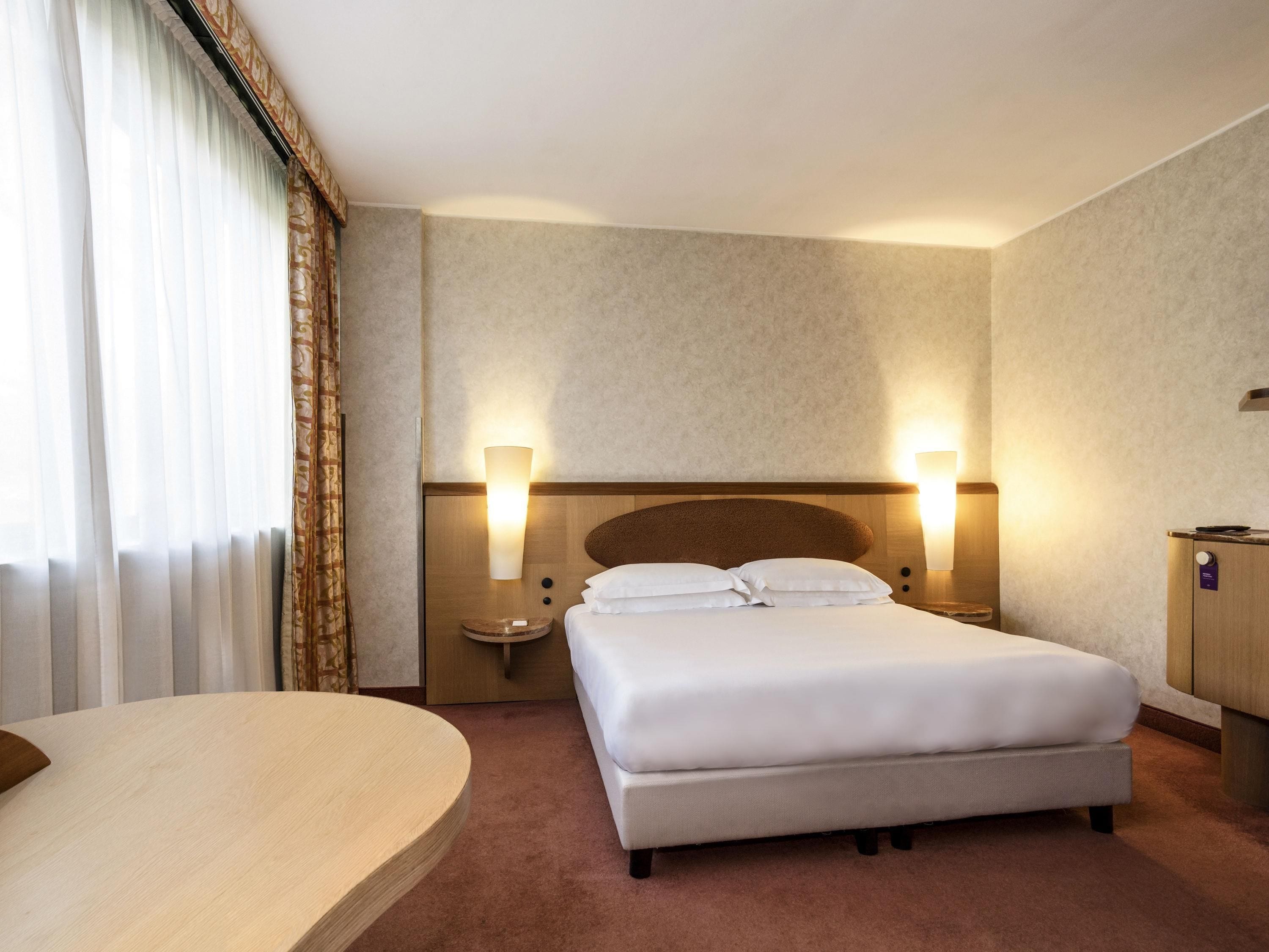 Une chambre d’hôtel avec un lit, deux lampes de chevet, une table de chevet, un bureau et une fenêtre avec des rideaux.