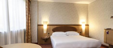 A hotel room with a bed, two bedside lamps, a nightstand, a desk, and a window with curtains.