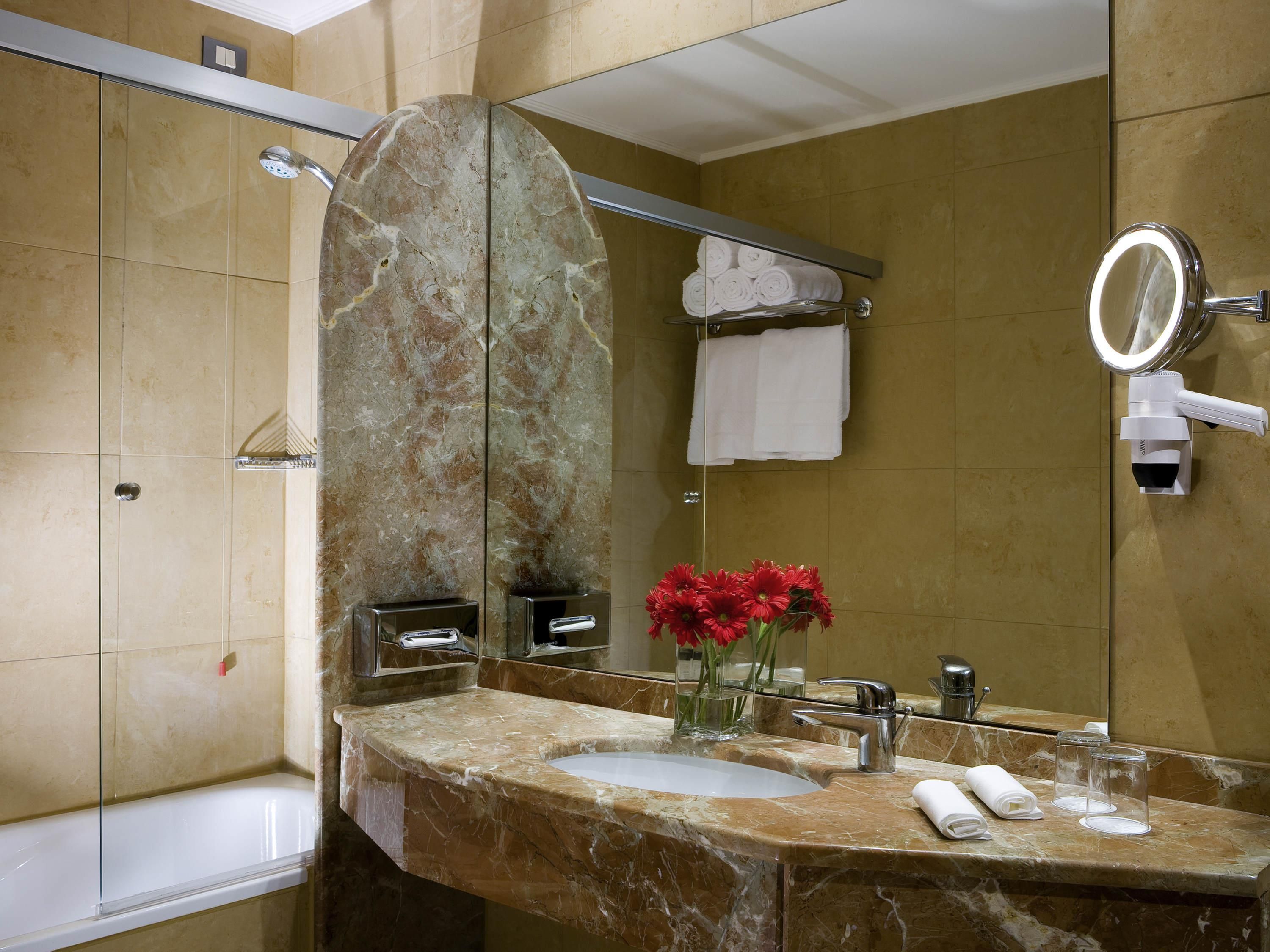 A bathroom with a marble sink, a glass shower enclosure, a towel rack with folded towels, a mirror with a light, and a vase with red flowers.