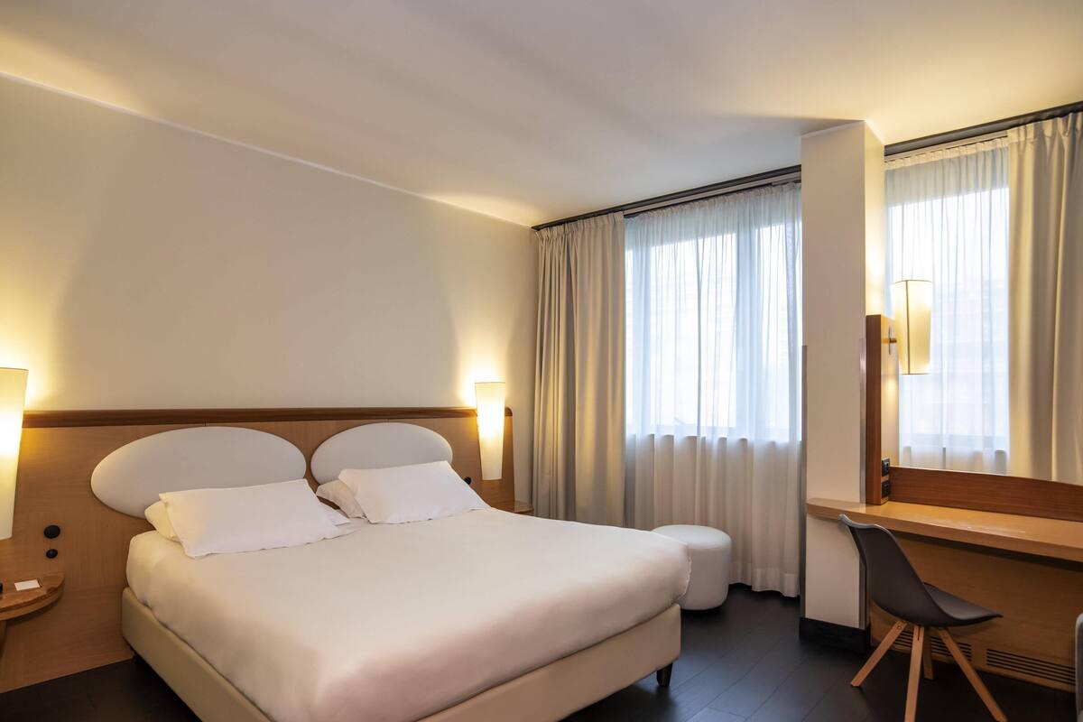 A hotel room with a large bed, bedside lamps, a desk with a chair, and a window with curtains.
