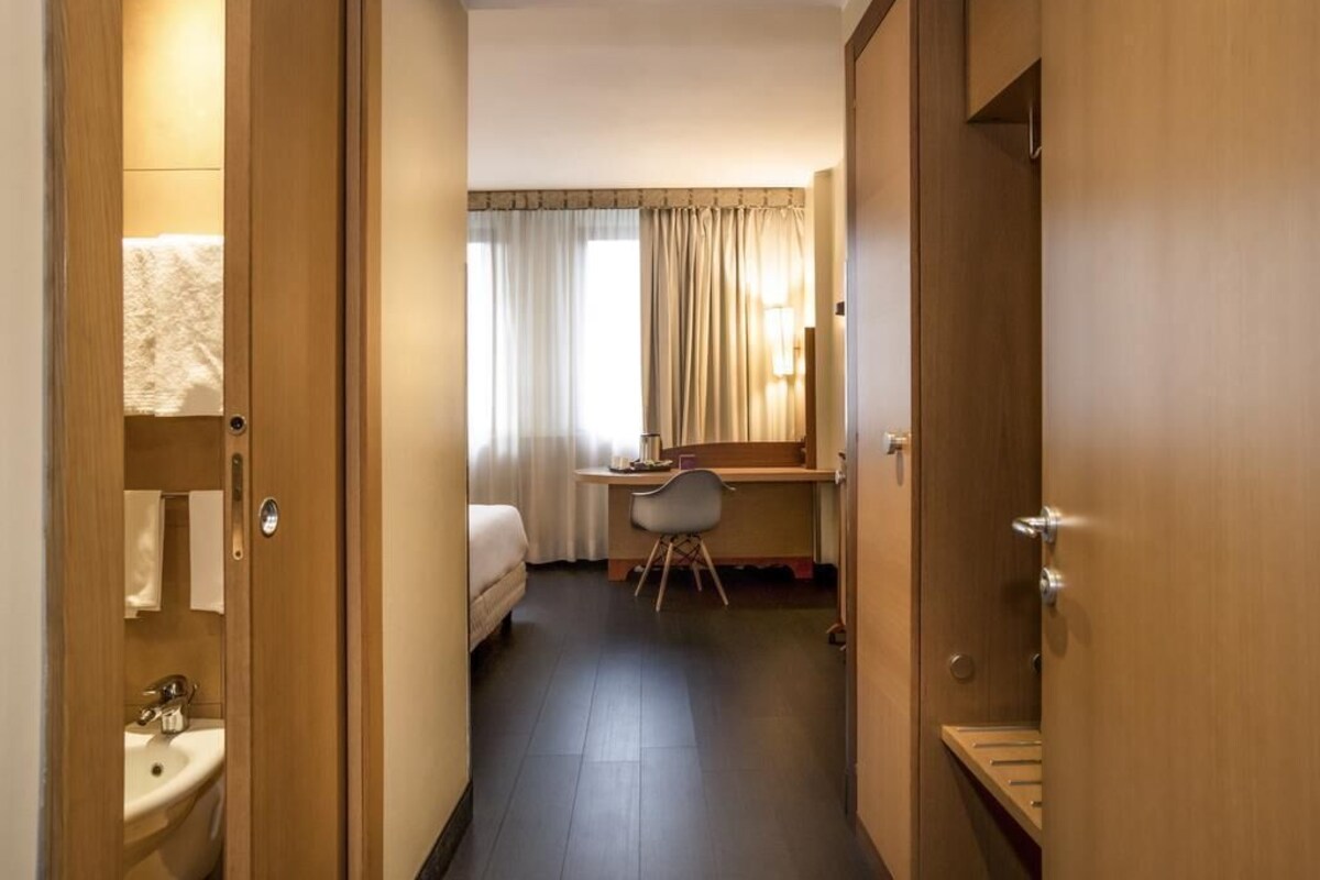 A hotel room with a bed, desk, chair, and a bathroom with a sink and toilet.