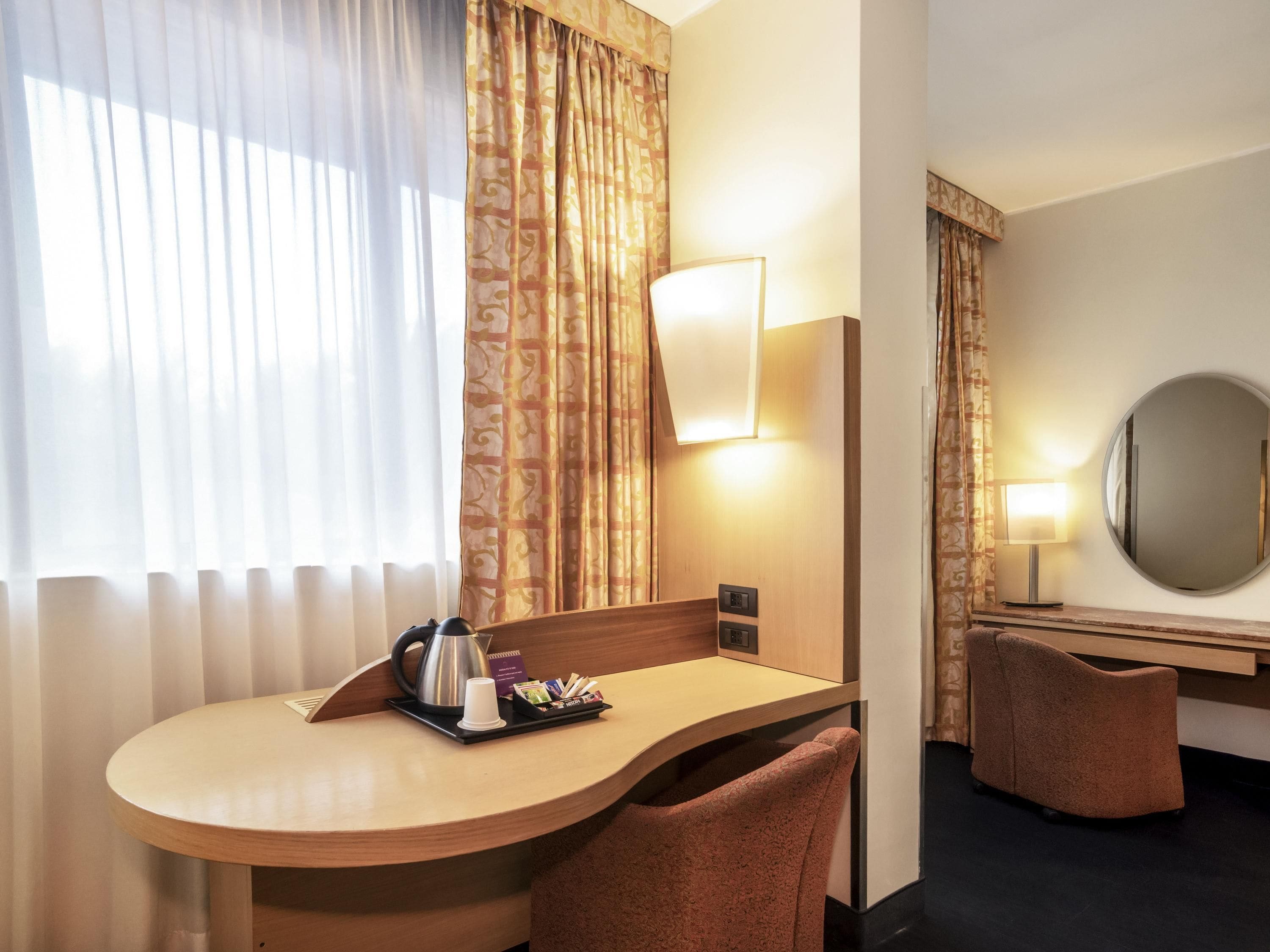 A hotel room with a curved wooden desk, a coffee pot, a tray with snacks, a mirror, and two chairs.