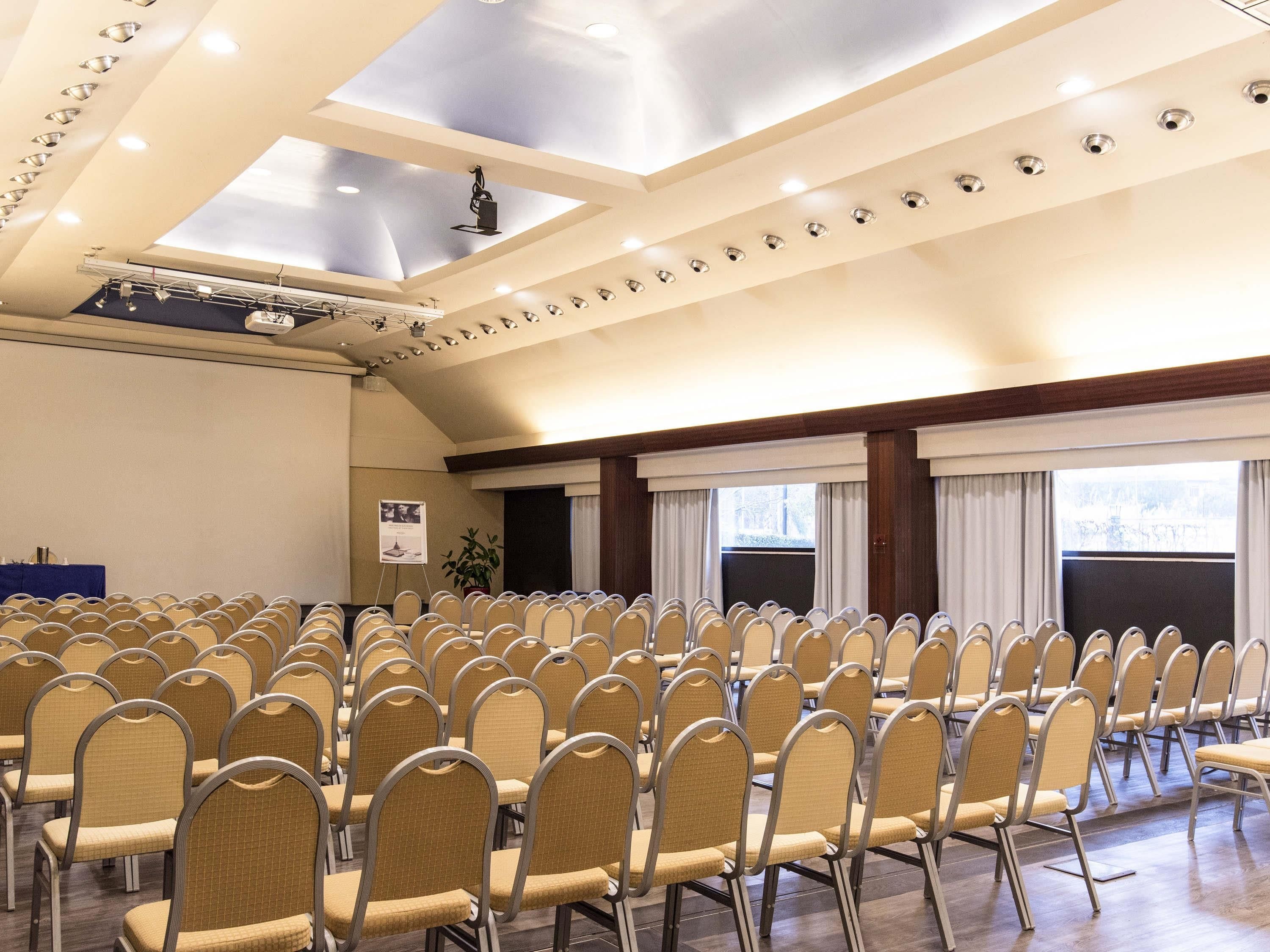 Un salón de conferencias con filas de sillas, un escenario con proyector y ventanas grandes con cortinas.