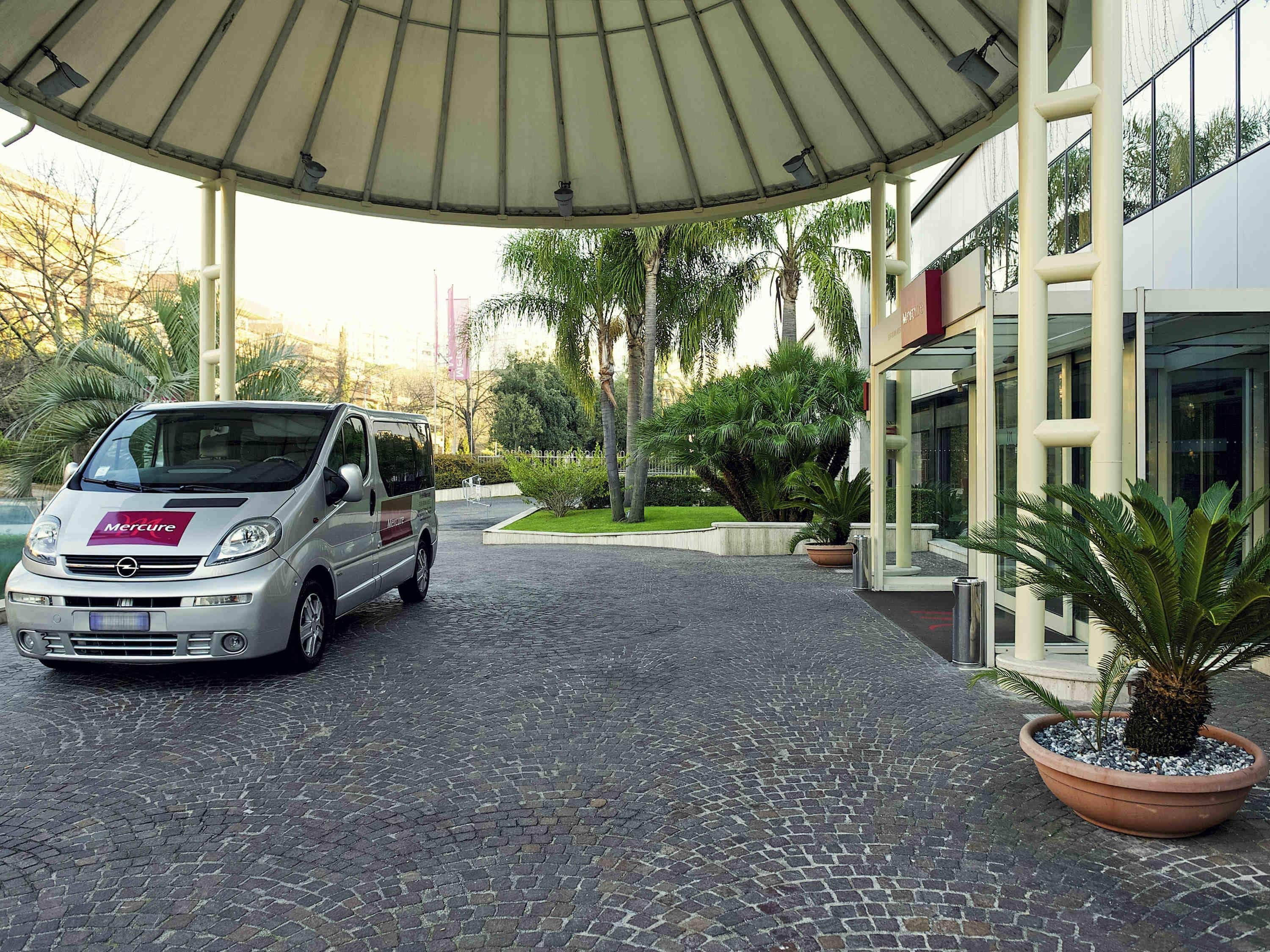 A silver van parked under a covered area with a paved walkway leading to a building with a modern facade.