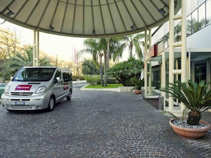 A silver van parked under a covered area with a paved walkway leading to a building with a modern facade. - Mercure Eur Roma West (Rome)