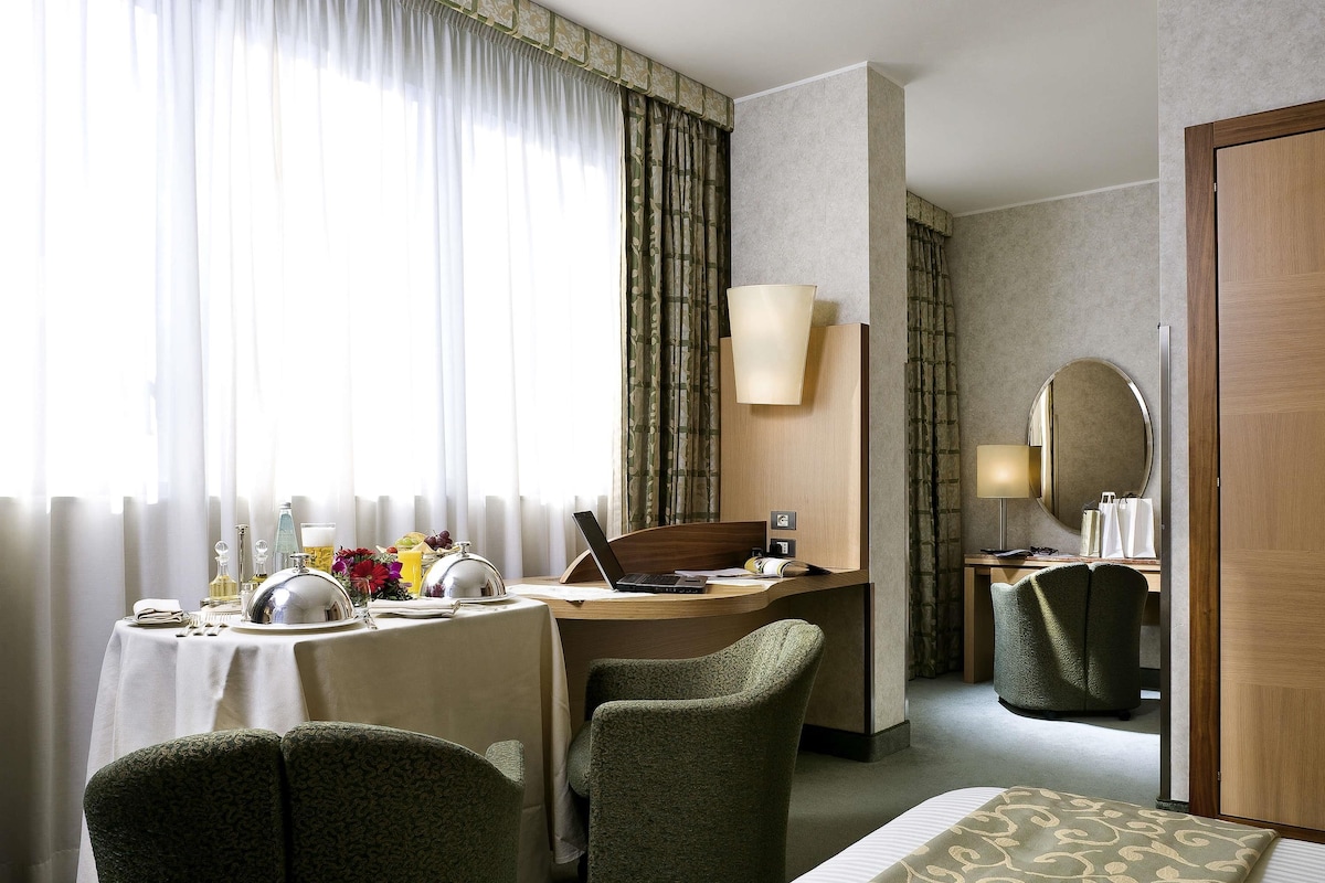 Een hotelkamer met een bed, een bureau met een laptop, twee stoelen, een eettafel gedekt voor twee personen en een groot raam met gordijnen.