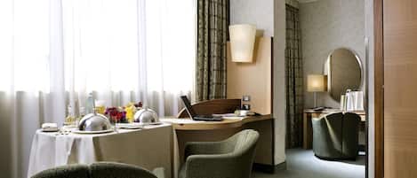 A hotel room with a bed, a desk with a laptop, two chairs, a dining table set for two, and a large window with curtains.