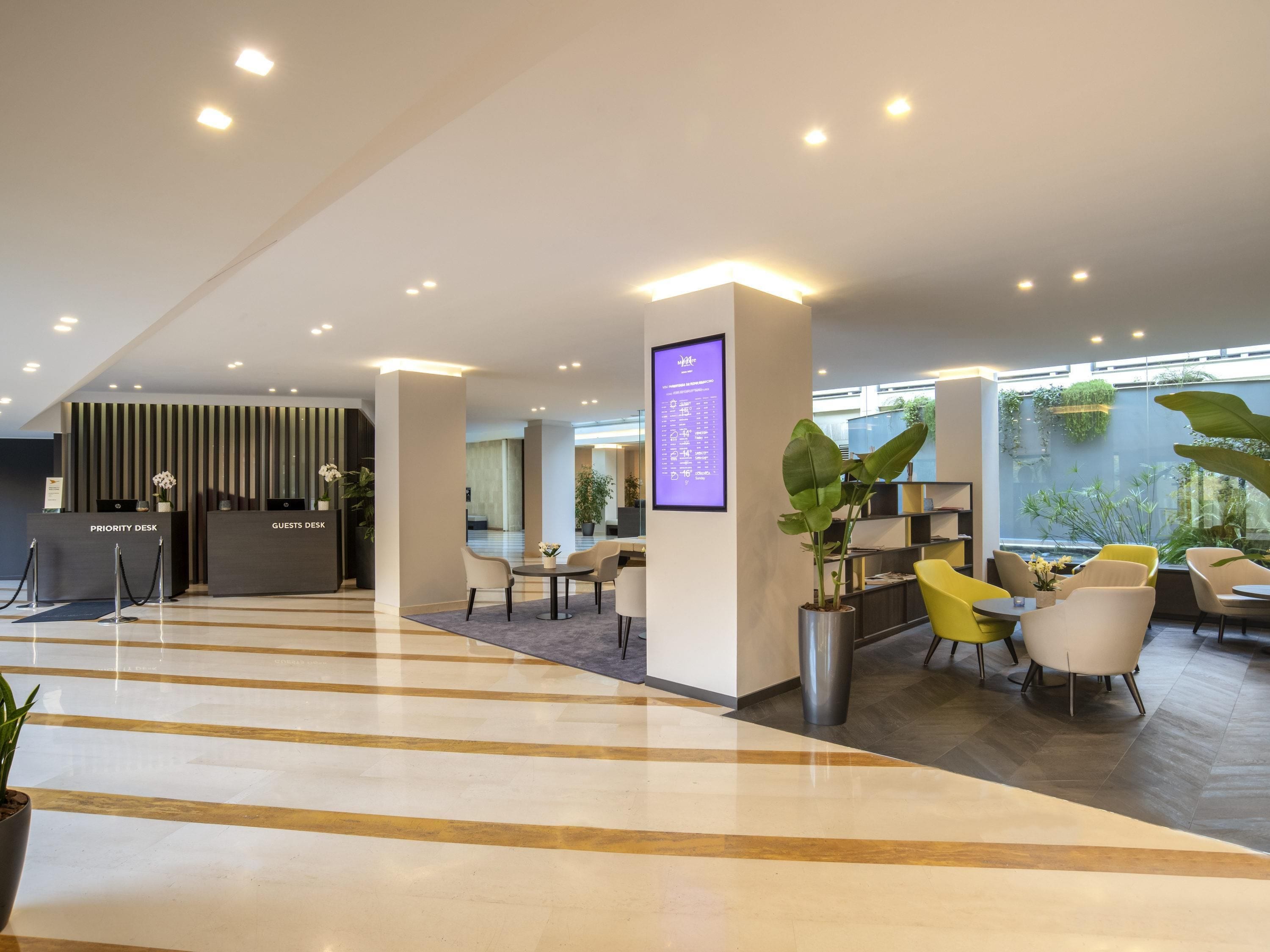 A modern lobby with priority and guest desks, seating areas, a digital menu board, and a large window with plants.