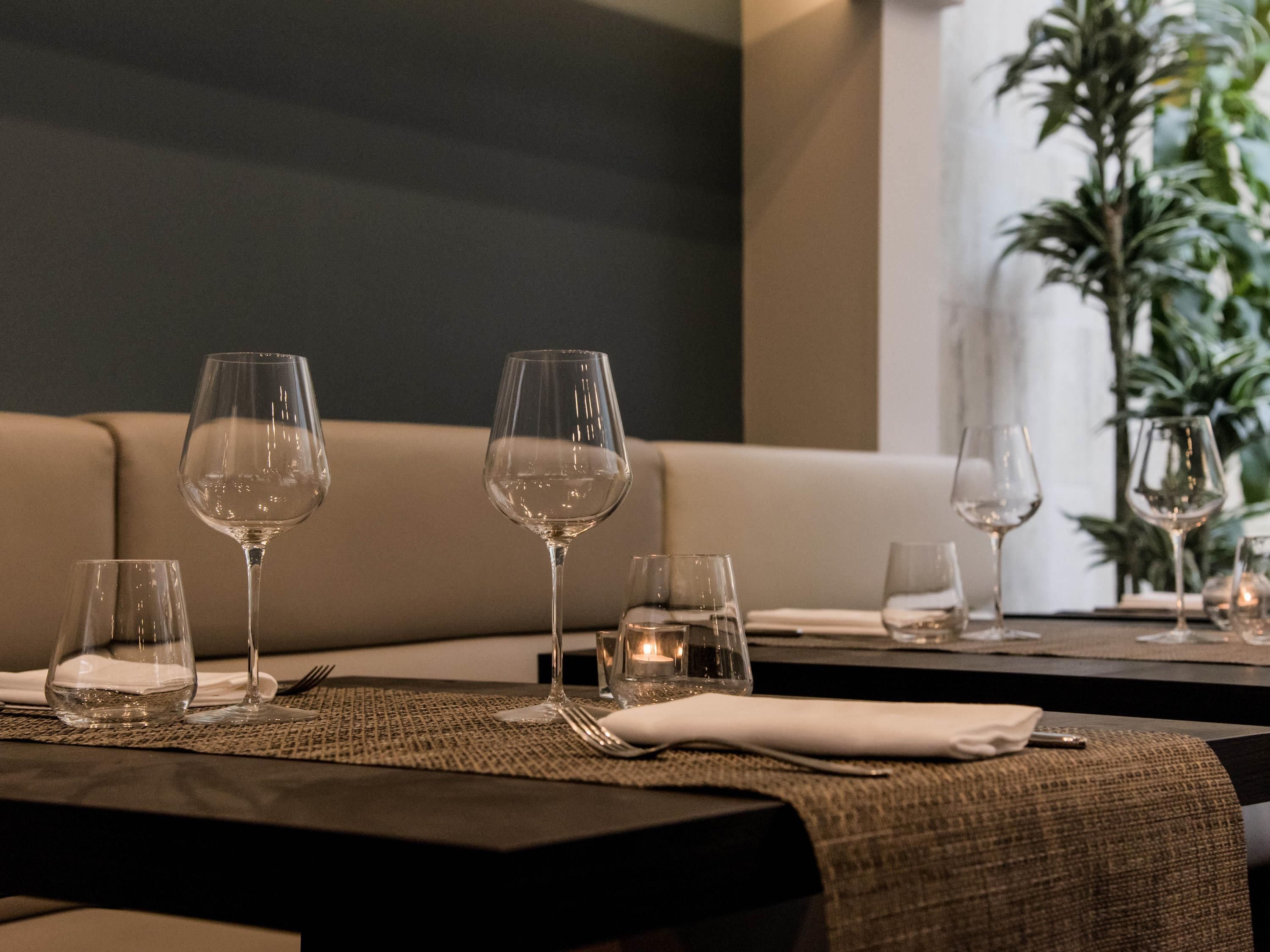Una mesa de comedor dispuesta con copas de vino, platos y cubiertos, con un sofá y una planta al fondo.