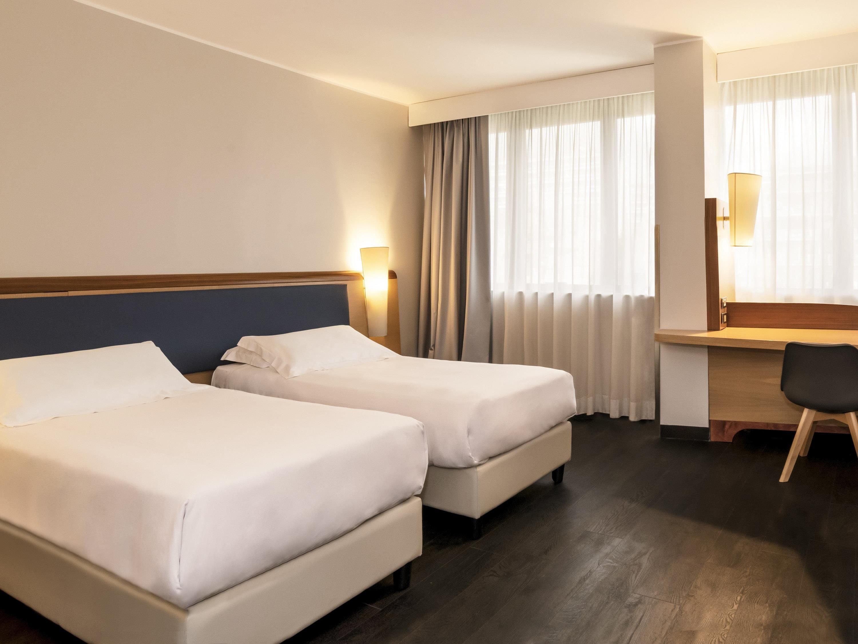 A hotel room with two beds, a desk, a chair, a lamp, and a window with curtains.