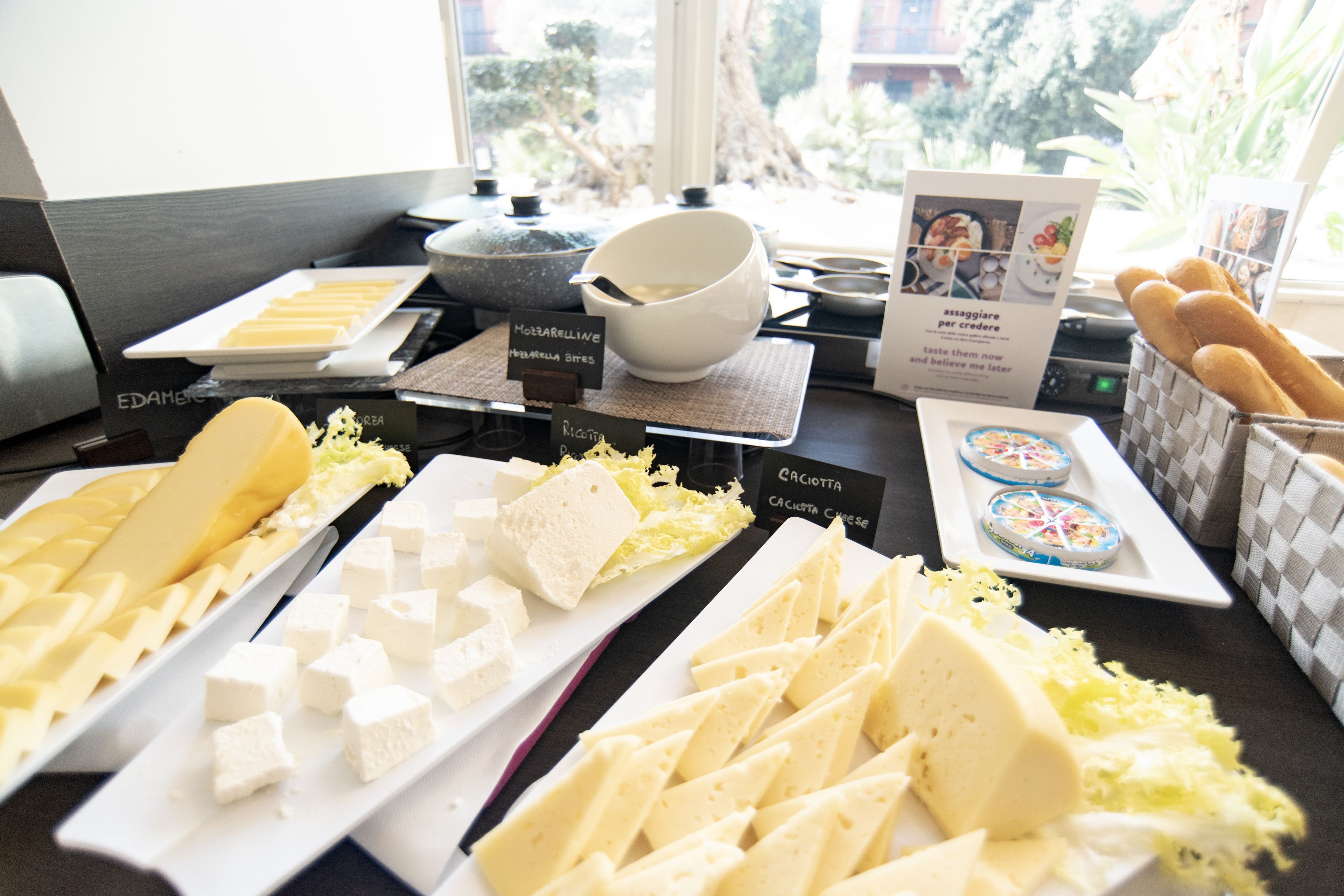 A buffet table with various cheeses, bread, and a bowl of soup.