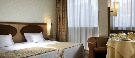 A hotel room with two beds, a sitting area with a table set for breakfast, and a large window with curtains.